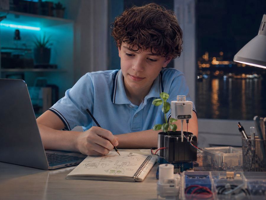 A teenager sketches a wiring diagram beside a basil seedling wired to a moisture sensor, a laptop open and trays of components across the desk. The hunch has been turned into a small, cheap test.